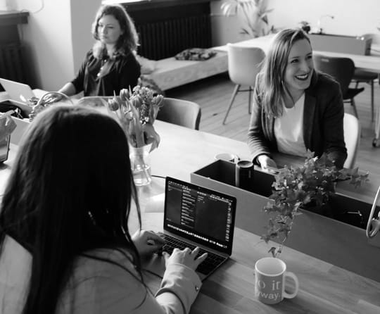 2 women working at a desk and one woman laughing at something off-screen